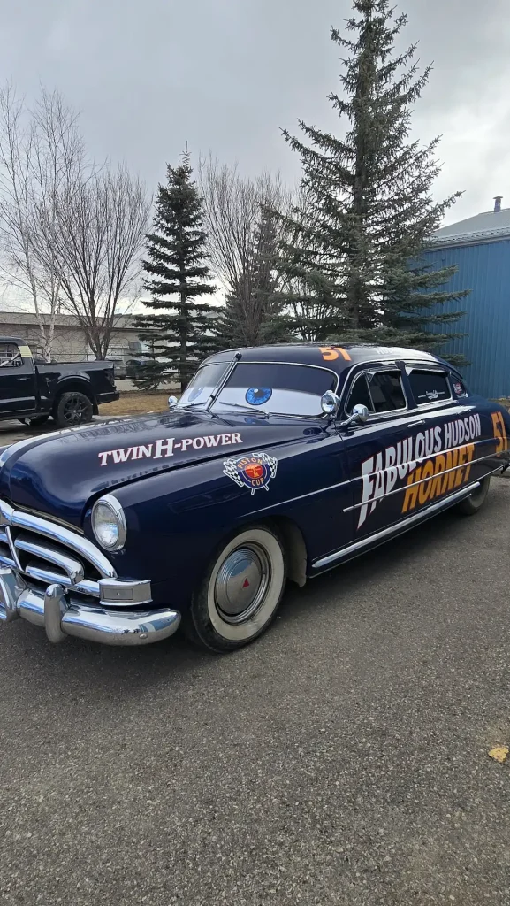 Custom stickers adorn a vintage blue car parked beside tall trees